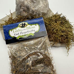Puede incluir: Una pequeña bolsa de plástico transparente llena de hierbas marrones secas y pequeñas semillas marrones. La bolsa tiene una etiqueta azul y negra con el texto "Lady of the Moss End Negativity" y una dirección de sitio web.