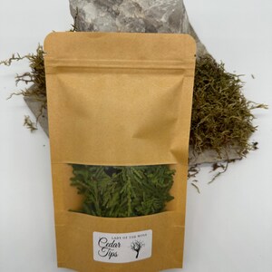 Może przedstawiać: Brązowa papierowa torba z przezroczystym okienkiem, wypełniona zielonymi końc&oacute;wkami cedru. Torba ma etykietę z napisem "Lady of the Moss Cedar Tips" z grafiką drzewa. Torba jest ustawiona na tle szarej skały i zielonego mchu.