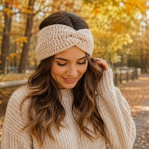 Op de afbeelding: Een beige gehaakte hoofdband met een centraal gedraaid detail, vervaardigd met een gestructureerd ribbelpatroon. Het accessoire is gemaakt van zacht garen en is ontworpen om rond het hoofd te worden gedragen om de oren te bedekken voor warmte.