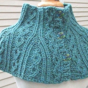 May include: A teal blue knitted cowl with a high collar and three decorative buttons. The buttons are circular and have a colorful swirling design.