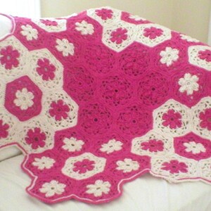 May include: A pink and white crocheted blanket with a floral pattern. The blanket is made of yarn and has a soft, textured surface.