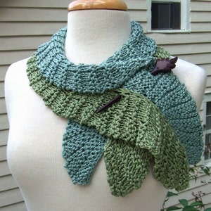 May include: A knitted scarf in shades of green and blue, with a wooden bird pin attached. The scarf is draped around a mannequin's neck.