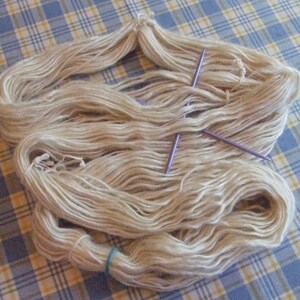 May include: A skein of natural white yarn with two purple knitting needles.