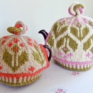 Könnte beinhalten: Zwei gestrickte Teekannenwärmer in einem grün-weiß-rosa Muster. Die Wärmer sind mit einem Kabelstrickmuster gearbeitet und haben oben einen dekorativen Knoten.