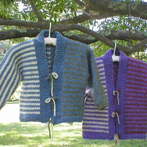Può includere: Due cardigan a righe di maglia con chiusura a cravatta. Il cardigan a sinistra è blu e verde con strisce bianche. Il cardigan a destra è viola e bianco con strisce bianche.