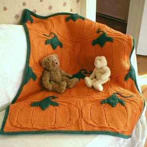 May include: A knitted orange and green pumpkin-themed baby blanket with two teddy bears sitting on it.