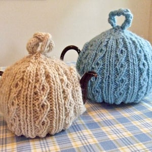 May include: Two knitted teapot cozies, one in beige and one in light blue, with a cable knit pattern. The cozies are sitting on a blue and white checkered tablecloth.