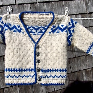 Può includere: Cardigan lavorato a maglia color crema con motivi geometrici blu e finiture. Il cardigan con bottoni è appeso a un filo per stendere i panni. Il design include uno scollo a V e maniche lunghe. Il cardigan è realizzato in filato morbido e strutturato.