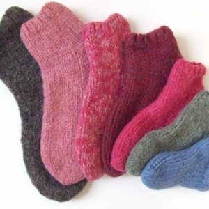 May include: A set of six knitted socks in various colours, including grey, pink, burgundy, and blue. The socks are all different sizes and are arranged in a row.