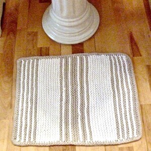May include: A rectangular, hand-knitted bath mat with vertical stripes in cream and beige. The mat is placed on a wooden floor, with a white pedestal sink base visible in the background.