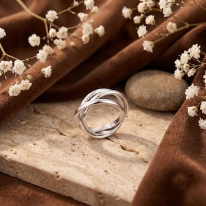 Può includere: Un anello color argento composto da tre fasce intrecciate che creano un design circolare continuo e sovrapposto. La superficie metallica è liscia e riflettente, caratterizzata da una finitura lucida che cattura la luce sulla struttura.