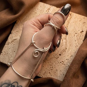 May include: A silver-toned metal hand harness featuring a smooth, continuous wire design that connects a wrist band to a ring worn on the thumb. The piece includes a decorative spiral element and a separate simple band ring on the index finger.