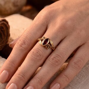 Può includere: Un anello color oro con una pietra preziosa rosso scuro centrale dal taglio a marquise. La fascia presenta dettagli in metallo lavorato, tra cui foglie scolpite e piccoli accenti floreali che fiancheggiano la pietra centrale su entrambi i lati.
