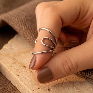 May include: A silver-toned metal ring featuring a multi-layered, open-ended design that wraps around the finger. The band consists of three distinct, smooth, polished wire-like loops that create a continuous, fluid, and stacked appearance.