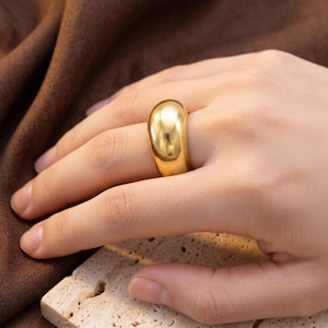 May include: A thick, polished gold-tone dome ring with a smooth, reflective surface. The ring features a wide, rounded band design that tapers slightly towards the palm side, showcasing a high-shine metallic finish that catches the light.