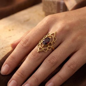 May include: A gold-toned ring featuring an elongated, openwork metal frame with a symmetrical petal-like design. A single, oval-shaped, deep red gemstone is securely set in the center of the decorative metal structure, which rests on a thin band.