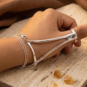 May include: A silver hand chain featuring a polished metal frame that encircles the thumb and connects to a delicate double-strand chain bracelet. The structure is rigid and triangular, extending from the thumb base to the wrist area.