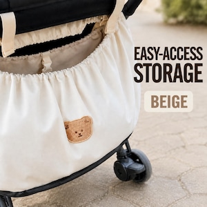 May include: A beige stroller storage bag with an elasticated opening and a small brown embroidered bear patch on the front. The text EASY-ACCESS STORAGE and BEIGE is printed on the right side of the image against a blurred outdoor background.
