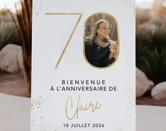 May include: A white sign with gold accents and the number 70. It includes a photo of a woman and the text "BIENVENUE À L'ANNIVERSAIRE DE Claire 18 JUILLET 2026". The sign is displayed on a wooden easel, suitable for a birthday celebration.
