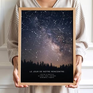 Peut inclure: Un tirage encadré représentant un ciel étoilé avec des constellations et la Voie lactée. L'impression présente un dégradé bleu foncé et violet, avec une silhouette de forêt en bas. Le texte en bas indique "LE JOUR DE NOTRE RENCONTRE" et comprend des noms, une date et des coordonnées.
