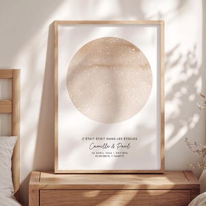 Peut inclure: Un cadre en bois clair encadre une impression avec une carte céleste beige. Le texte "C'ÉTAIT ÉCRIT DANS LES ÉTOILES" et les noms "Camille & Paul" sont présents, ainsi qu'une date et des coordonnées.