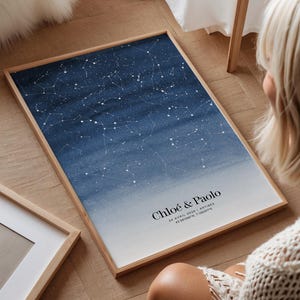 Könnte beinhalten: Ein gerahmter Druck mit einer aquarellartigen Darstellung eines Sternenhimmels in Blautönen. Der Druck enthält die Namen "Chloé & Paolo" sowie Datum und Ortsangaben. Der Rahmen ist aus hellem Holz.