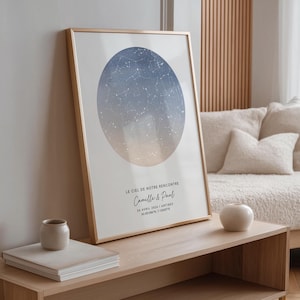 Peut inclure: Un cadre doré contenant une impression d'une carte céleste. L'œuvre présente un cercle dégradé bleu et blanc avec des constellations. Le texte en bas indique "LE CIEL DE NOTRE RENCONTRE" et des noms et une date. Un petit vase en céramique et des livres sont sur une table en bois.