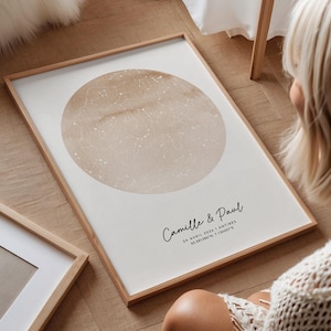 Può includere: Una stampa incorniciata con un design a mappa celeste. L'opera d'arte presenta un design circolare beige con motivi di stelle bianche su sfondo bianco. La cornice è in legno chiaro. I nomi "Camille & Paul" sono scritti in corsivo, insieme a una data e alle coordinate.