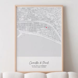 Peut inclure: Une impression encadrée d'une carte de ville détaillée en niveaux de gris, avec un cœur rouge marquant un lieu spécifique. L'impression comprend les noms "Camille & Paul" et le texte "LA OÙ TOUT A COMMENCÉ 18 AVRIL 2026 | CANNES" en écriture élégante.
