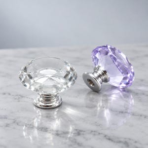 Puede incluir: Dos pomos para armario facetados con forma de diamante. Uno es transparente e incoloro, mientras que el otro tiene un tono violeta claro. Ambos pomos cuentan con una base de metal plateado pulido con un orificio roscado para su montaje.