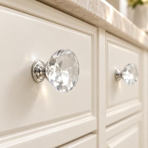 May include: A clear, faceted, diamond-shaped glass cabinet knob with a polished chrome metal base. The knob is mounted onto a cream-colored wooden drawer front with a recessed panel design, reflecting light through its multi-faceted surface.