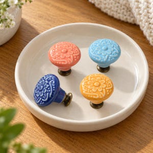 Puede incluir: Cuatro pomos redondos de cerámica para armarios con patrones florales en relieve en color coral, azul claro, azul oscuro y amarillo. Cada pomo presenta un diseño floral central rodeado de volutas y una base de metal bronce oscuro.