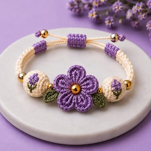 May include: A macrame bracelet featuring a central purple crocheted flower with a gold bead center, flanked by two cream-colored beads embroidered with lavender sprigs and green leaves, accented with gold beads and a sliding knot closure.