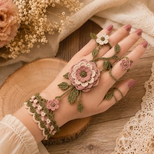 May include: A crochet hand flower bracelet featuring a large layered pink and cream flower centerpiece with green leaves, connected by delicate green vines to a matching ring and a scalloped wrist cuff adorned with small pink and cream flowers.