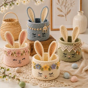 May include: Four round crochet baskets featuring bunny ears and embroidered faces. The baskets are pink, blue, cream, and green, decorated with crocheted flowers or lace trim. A natural woven basket and a white vase are also visible in the scene.