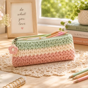 Puede incluir: Un estuche rectangular de ganchillo con rayas horizontales en verde salvia, crema y rosa empolvado. El estuche tiene una cremallera metálica con un pequeño colgante de flor de ganchillo a juego en el tirador. Se ven varios lápices de colores dentro.