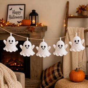 Puede incluir: Una guirnalda con cinco adornos de fantasmas blancos tejidos a ganchillo y colgados de un cordel marrón. Cada fantasma tiene una cabeza redondeada, brazos extendidos, un borde inferior festoneado y ojos y boca sonriente bordados en negro.