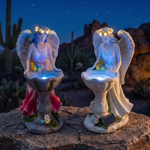 May include: Two angel statues with illuminated floral crowns and wings, each holding a birdbath basin that glows with a blue light. The left angel wears a pink dress, while the right angel wears a white dress. Both feature solar panels at the base.