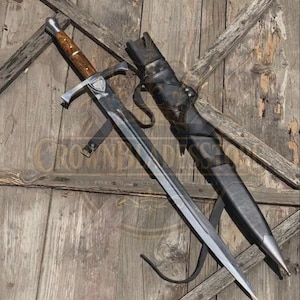 Puede incluir: Una espada de acero con pomo de metal pulido, empuñadura de madera veteada marrón y guarda plateada. La hoja es de doble filo y termina en punta. Junto a la espada hay una vaina de cuero negro con una punta de metal plateado y correas.
