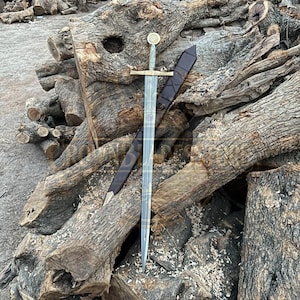 Pode incluir: Uma espada longa de aço com guarda metálica e pomo circular com um design gravado. A espada é acompanhada por uma bainha de couro castanho escuro com ponta afiada, ambas apoiadas contra uma pilha de troncos de madeira rugosos.
