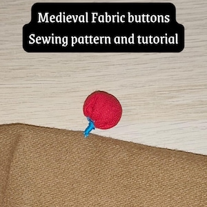 Può includere: Un bottone in tessuto rosso fissato su un pezzo di tessuto marrone chiaro con filo blu. Il testo "Medieval Fabric buttons Sewing pattern and tutorial" è visualizzato in un riquadro nero nella parte superiore dell'immagine.