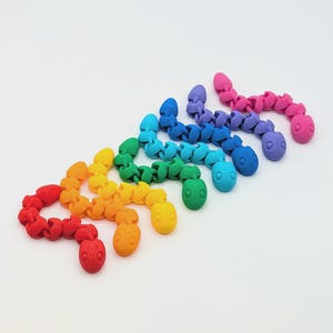 May include: A collection of seven articulated snake toys arranged in a diagonal row, featuring a vibrant rainbow colour gradient from red, orange, yellow, green, light blue, dark blue, to purple and pink, each with a textured, segmented body.