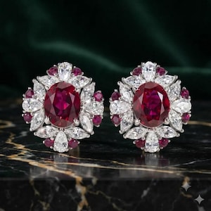 Puede incluir: Un par de pendientes de botón con una gran piedra central ovalada de color rojo intenso rodeada por un halo de piedras transparentes de talla marquesa y piedras rojas más pequeñas de talla redonda en una montura de metal plateado.