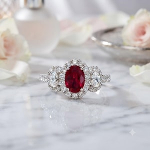 Puede incluir: Un anillo de tono plateado con un gran rubí rojo intenso de corte ovalado en el centro, rodeado por un halo de pequeños diamantes blancos transparentes. Dos diamantes blancos ovalados adicionales están engastados a cada lado del rubí central.