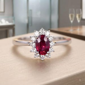 Puede incluir: Un anillo de tono plateado con un rubí central de color rojo intenso en talla oval, rodeado por un halo de doce diamantes pequeños, redondos y blancos transparentes. La banda es lisa y pulida, con un diseño clásico de engaste floral.