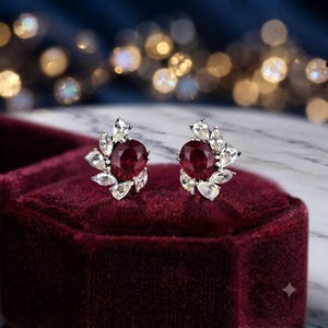 Könnte beinhalten: Ein Paar Ohrstecker mit einem zentralen, oval geschliffenen, tiefroten Edelstein, der von einer Anordnung aus marquise-geschliffenen, klaren weißen Steinen in einer metallischen Fassung in einem floralen Design umgeben ist.