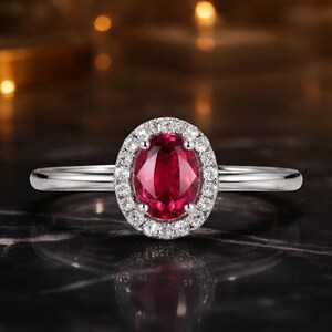 Puede incluir: Un anillo de tono plateado con una piedra preciosa de color rojo intenso de corte ovalado en el centro, rodeada por un halo de pequeñas piedras claras de corte redondo. La banda es lisa y pulida, con un engaste clásico de garras.