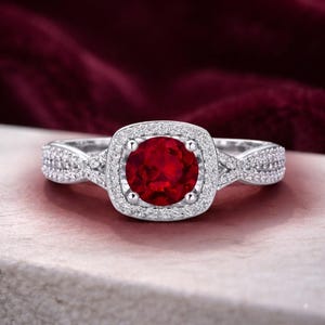 Op de afbeelding: Een zilverkleurige ring met een ronde, dieprode middensteen omringd door een halo van kleine, heldere pavé-gezette steentjes. De band is verdeeld in twee in elkaar grijpende strengen, elk ingelegd met piepkleine heldere steentjes voor een sprankelend effect.
