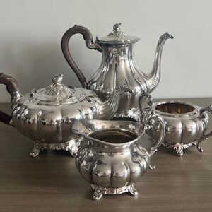 May include: A silver tea set consisting of a tall teapot, a shorter teapot, a creamer, and a sugar bowl. Each piece features a fluted, scalloped body, ornate feet, and dark brown handles. The teapots have lids with small finials and curved spouts.