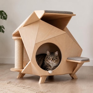 May include: A geometric cat house made of light-colored wood featuring a circular entrance, a sisal-wrapped scratching post, and two gray fabric-covered platforms. A brown tabby cat is resting inside the central circular opening of the structure.